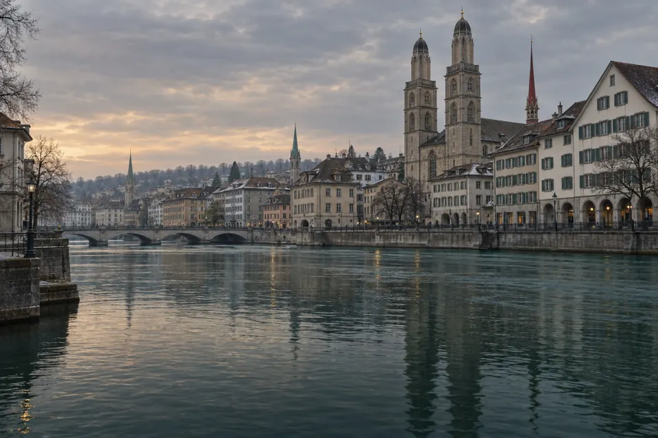 The Limmat river through Zürich today, old-town rooftops and church towers along the bank, cold green water under a low grey winter morning sky, the quay empty and calm