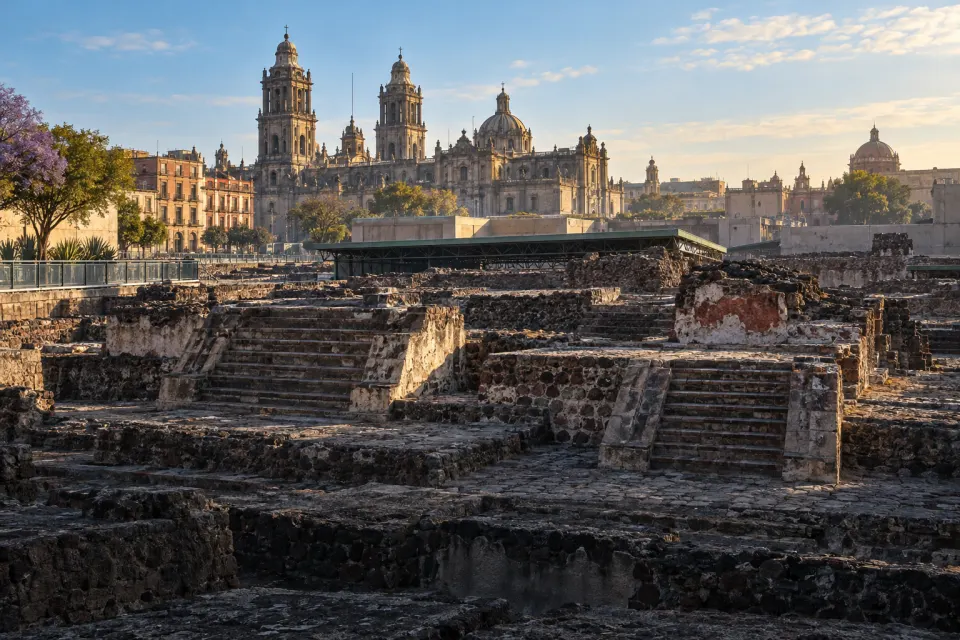The excavated ruins of the Templo Mayor in Mexico City, twin stepped bases and the round carved Coyolxauhqui stone in the foreground, cathedral rooftops beyond, high-altitude morning light