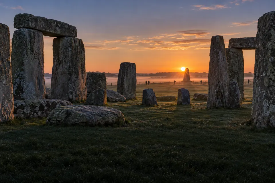 Stonehenge's sarsen circle and trilithons on Salisbury Plain with the midsummer sun rising over the Heel Stone, ground mist, a few visitors in modern clothes at a distance, cold clear dawn