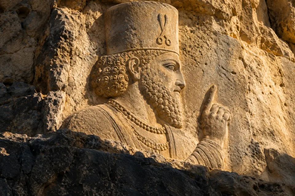 Kartir as a Sasanian rock relief at Naqsh-e Rajab, a bearded priest in profile in a tall cylindrical cap with one hand raised, carved on a Fars cliff, hard noon sun